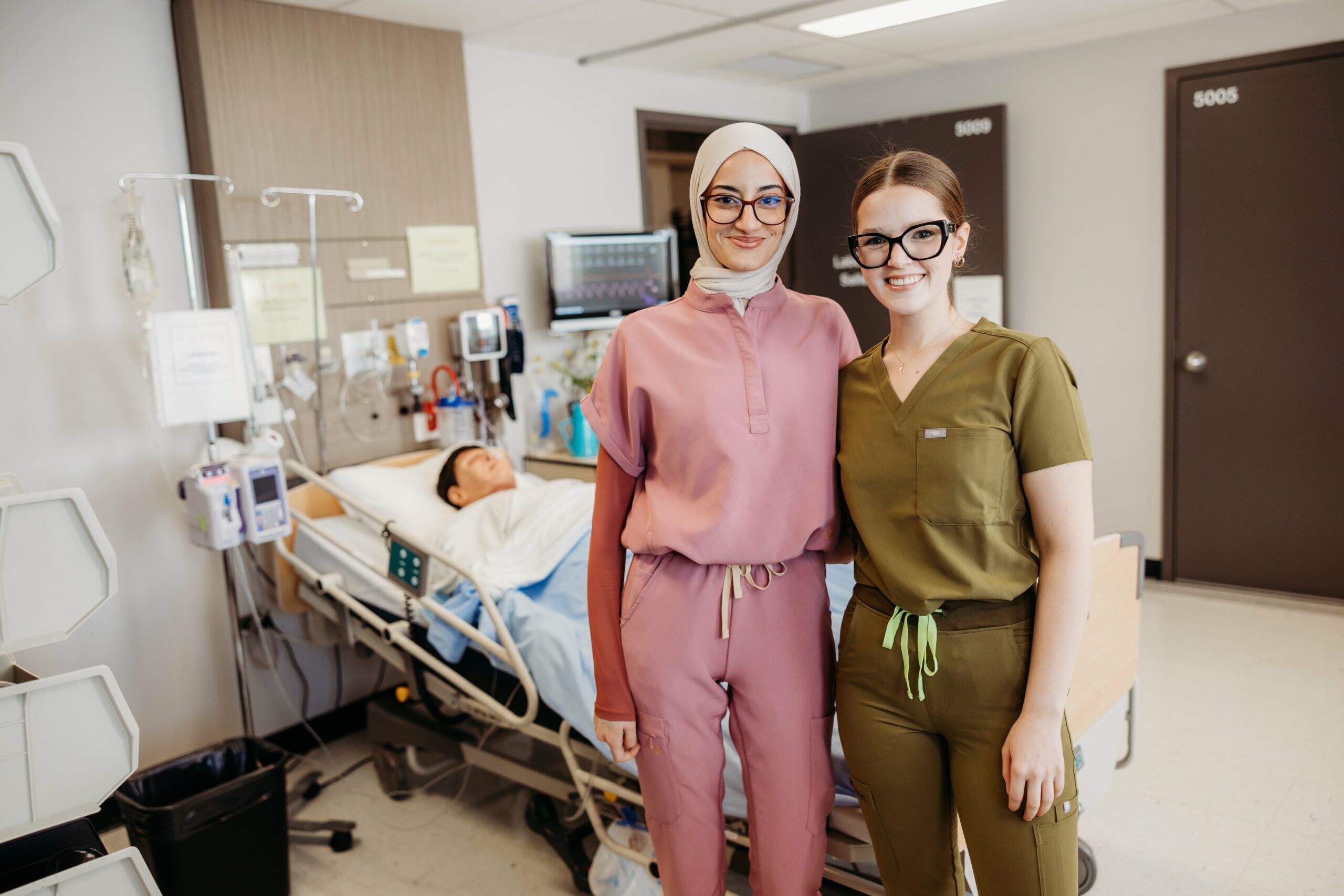 Deux étudiantes en soins infirmiers sourient à la caméra dans une salle avec civière et installations médicales.